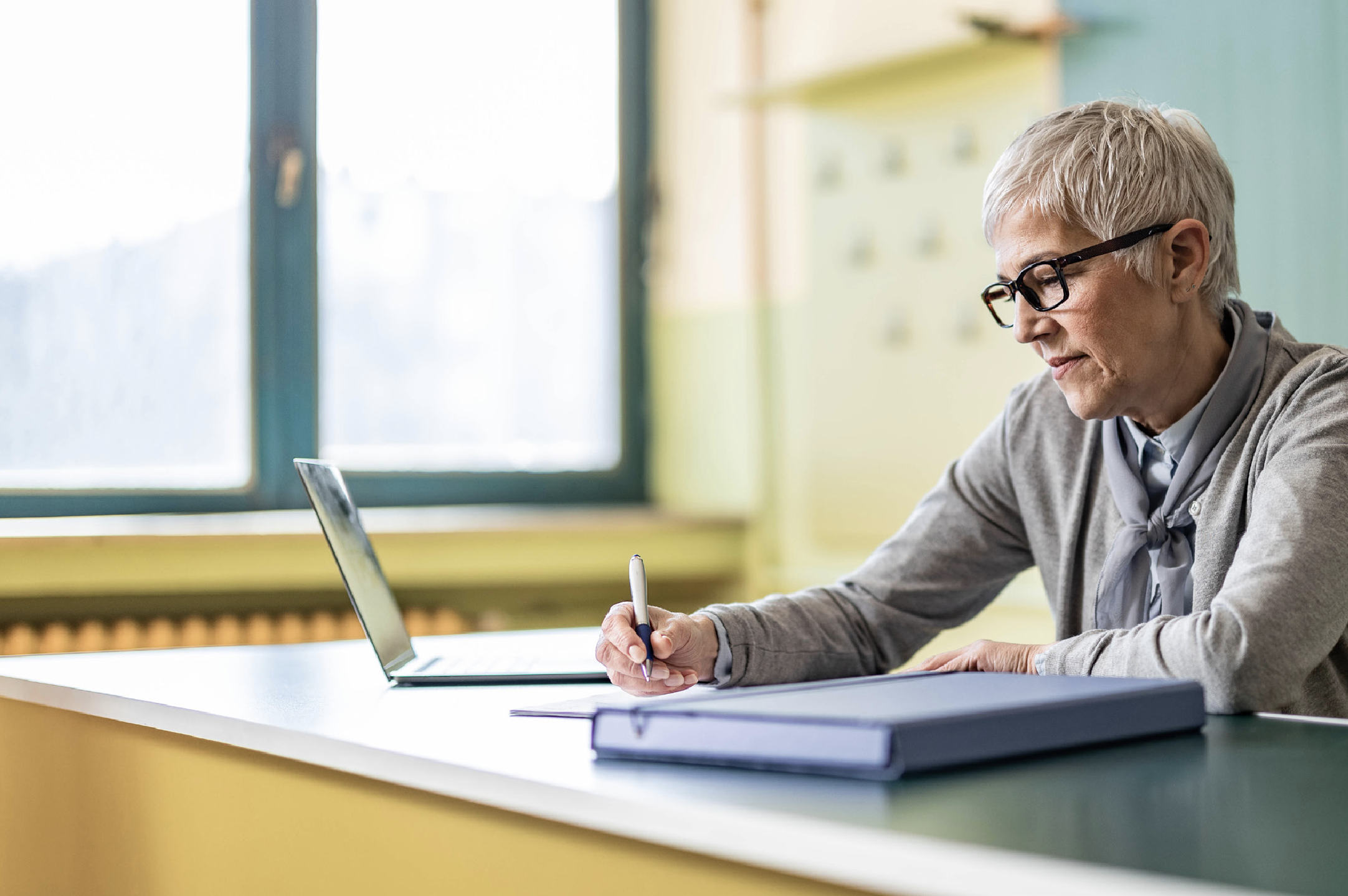 Photo Of Teacher Wearing Glasses Writing At Desk With Laptop@2x
